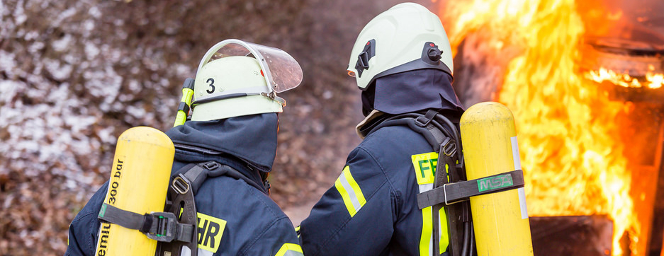 2 Feuerwehrmänner vor Flammen, die ein Auto löschen